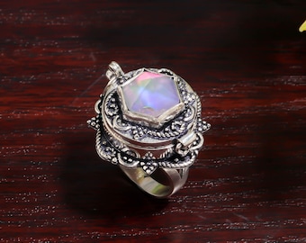 Puede incluir: Anillo de plata ornamentado con una gran gema hexagonal iridiscente. El anillo tiene filigrana detallada y un compartimento con bisagras. La gema muestra un espectro de colores. El anillo está sobre una superficie de madera oscura.