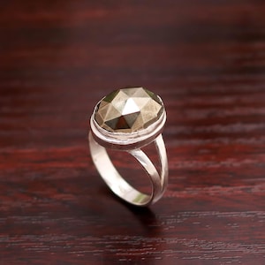 Pode incluir: Anel de prata com uma pedra preciosa oval facetada de cor castanho-dourado metálico. O anel tem um design de haste dividida. Está colocado sobre uma superfície de madeira castanho-avermelhada escura.