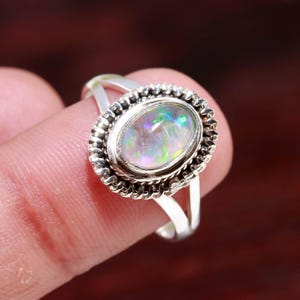 May include: A silver ring with an oval opal gemstone displaying iridescent colours. The opal is set in a detailed, textured bezel. The ring has a split shank design.