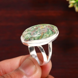 Peut inclure: Bague en argent ornée d'une pierre ovale verte et rouge. La pierre est sertie dans un anneau en argent poli avec une monture fendue. La bague est présentée sur un fond marron foncé.