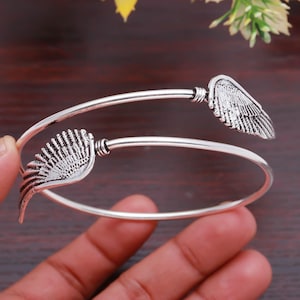 May include: A silver cuff bracelet with wing motifs at each end. The wings are intricately detailed with etched feather patterns. The open design allows for adjustment. The bracelet is displayed against a dark wooden background.