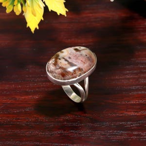May include: Oval gemstone ring with a silver band. The stone features a mix of brown, pink, and white hues. The ring's design includes a split shank, adding a unique touch to the jewelry.
