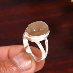 May include: A silver ring featuring a large, oval-shaped, light brown gemstone. The gemstone is set in a bezel setting, and the ring has a split shank design. The ring is held by a person's hand.