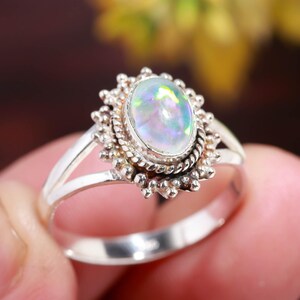 May include: A silver ring featuring an oval opal gemstone with iridescent colors. The gemstone is set in a detailed, ornate setting with a rope-like border and a sunburst design. The ring has a split shank.