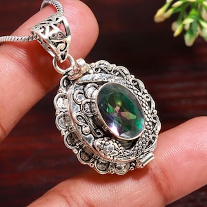 May include: Silver pendant necklace featuring an oval, iridescent gemstone. The pendant has an ornate, filigree design with a decorative border. The necklace is held by a hand.