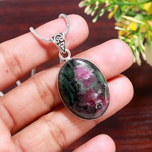 May include: Oval pendant necklace with a large, polished gemstone in deep red and dark green. The pendant is set in silver-coloured metal and hangs from a delicate chain. The gemstone is approximately 3.8 cm long.