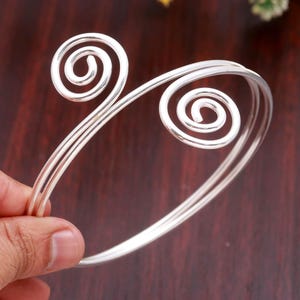May include: A silver-toned metal cuff bracelet featuring a double-wire design that terminates in two prominent, circular spiral motifs at each open end. The bracelet has a smooth, polished finish and a flexible, open-ended construction.