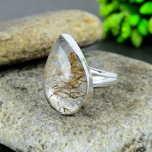 Op de afbeelding: Een zilveren ring met een druppelvormige, heldere edelsteen met bruine en gouden insluitsels. De ring heeft een gesplitste band en een gepolijste zilveren bezel. De ring wordt gepresenteerd op een grijze stenen ondergrond.