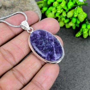 Può includere: Una collana con pendente in argento con una pietra preziosa ovale, viola scuro. La pietra è incastonata in una lunetta d'argento e pende da una sottile catena d'argento. La gemma ha un aspetto strutturato, con sfumature di viola più chiare e più scure.