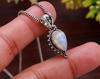 May include: A silver pendant necklace featuring a pear-shaped, iridescent moonstone. The pendant is intricately detailed with small beaded accents and a decorative bail. The chain is a delicate, textured design. The necklace is held by a hand.