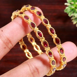 Puede incluir: Dos brazaletes dorados con piedras preciosas ovaladas de color rojo intenso. Los brazaletes se muestran, destacando el diseño detallado y el contraste entre el oro y las piedras rojas. Joyas para moda u ocasiones especiales.