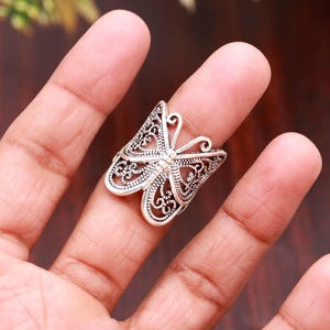 May include: A silver-toned ring featuring a butterfly design with intricate filigree cutouts. The metal surface has a darkened, oxidized finish within the recessed areas of the wings, highlighting the detailed scrollwork patterns throughout.