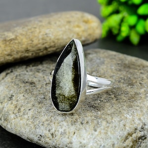Puede incluir: Un anillo de plata con una gran piedra preciosa en forma de lágrima. La piedra preciosa tiene una superficie oscura y reflectante con inclusiones doradas y verdes. La banda del anillo está dividida, agregando un elemento de diseño único.