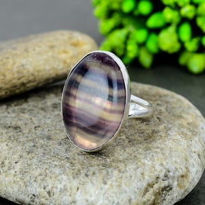 May include: A silver ring with a large, oval gemstone in shades of purple, lavender, and cream. The stone is set in a polished silver band. The ring is presented on a textured, grey stone. The ring's design is elegant and eye-catching.
