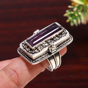 Könnte beinhalten: Ein rechteckiger Silberring mit einem großen, tiefvioletten Edelstein. Der Ring hat ein verziertes Filigranmuster um den Stein und entlang des Bandes. Der Edelstein hat eine tiefe, satte violette Farbe.