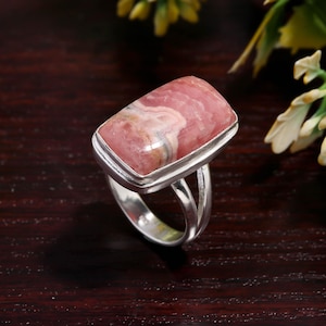 May include: A rectangular rhodochrosite gemstone ring. The ring features a polished, pink and white stone set in a silver band. The stone has a rectangular shape with rounded corners and a marbled appearance. The ring's band is split into two parallel strands.
