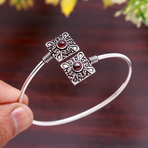 May include: A silver-toned cuff bracelet featuring two square, ornate settings. Each setting has a central, round, red gemstone. The bracelet's design includes intricate patterns and a simple, curved band.