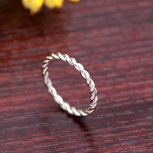 May include: A silver-colored ring with a twisted rope design. The ring is thin and delicate, with a continuous pattern of interwoven strands. The ring is set against a dark, reddish-brown background.
