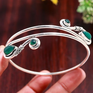 May include: A silver cuff bracelet featuring two parallel bands that curve into open ends. Each end is decorated with a smooth, dark green teardrop-shaped stone and a smaller round green stone, accented by intricate silver wire wrapping details.