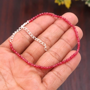 May include: A delicate bracelet featuring small, round, red beads strung together. The bracelet has a silver-colored chain with an adjustable clasp. The beads are a vibrant shade of red, creating a simple yet elegant accessory.