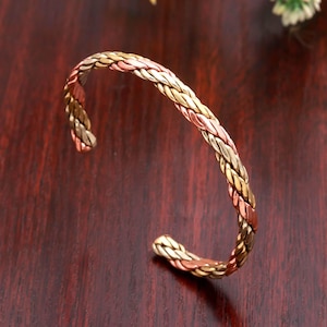 Può includere: Un bracciale a fascia composto da tre fili metallici intrecciati in tonalità alternate di rame, ottone e oro. Il bracciale presenta una texture ritorta simile a una corda lungo tutto il design circolare, con un'apertura sul retro.