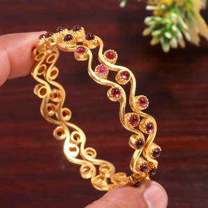 May include: A gold-coloured bangle with a wavy design, decorated with small, round, red gemstones. The bangle is held in a hand, highlighting its intricate details and polished finish. The gemstones are evenly spaced along the wavy pattern.