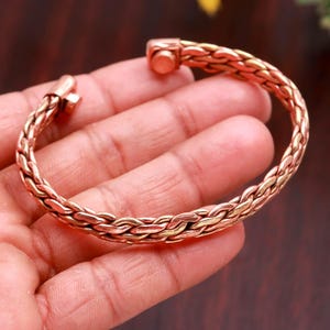 May include: A copper-toned metal cuff bracelet featuring a braided, woven texture throughout the band. The bracelet has a circular opening and terminates at both ends with small, cylindrical magnetic caps that face each other.