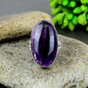 Könnte beinhalten: Ein Silberring mit einem großen, ovalen Amethyst-Edelstein. Der Amethyst hat eine tiefviolette Farbe mit polierter Oberfläche. Der Ring liegt auf einer hellen Steinoberfläche.