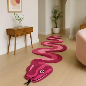 May include: A vibrant, snake-shaped rug in shades of pink and magenta. The rug features a detailed snake head with an eye and a forked tongue. The rug is placed on a light-colored wooden floor.