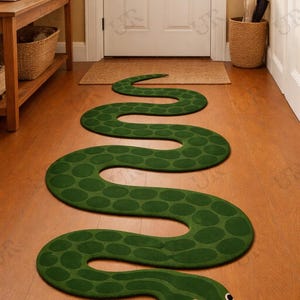 Può includere: Un tappeto verde a forma di serpente con un corpo a motivi e una testa dettagliata con occhi neri e una lingua biforcuta. Il tappeto è posizionato su un pavimento in legno in un corridoio, creando un ingresso stravagante.