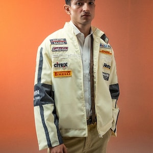Peut inclure: Veste de course crème avec des accents noirs et gris, ornée d'écussons de sponsors tels que "Citrix" et "Hertz". La veste zippée est portée sur une chemise blanche à col et un pantalon kaki. Le style général est sportif et à la mode.