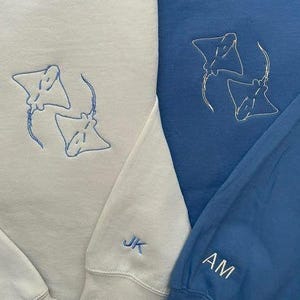 May include: Two crewneck sweatshirts, one cream and one blue, are displayed. Each sweatshirt features an embroidered design of two manta rays. The cream sweatshirt has "JK" embroidered on the sleeve, while the blue one has "AM".