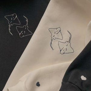 May include: Three sweatshirts in black and cream colors. Each sweatshirt features an embroidered design of two manta rays. The cuffs of the sleeves have a small heart embroidered on them. The sweatshirts are made of a soft material.