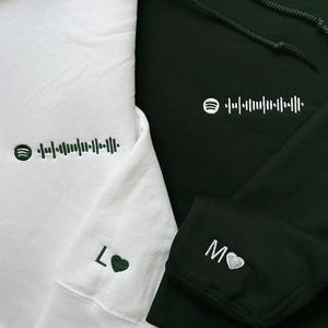 May include: Two hooded sweatshirts, one white and one dark green, are displayed. Each has a Spotify code and a small embroidered design. The white sweatshirt has "L" and a heart on the sleeve. The dark green sweatshirt has "M" and a heart on the sleeve.