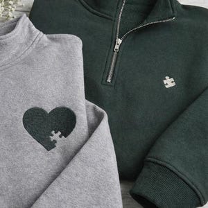 以下が含まれることがあります： 2つのクォータージップスウェットシャツ。1つは、濃い緑色のハート型のパズルピースのデザインが施されたヘザーグレーです。もう1つは、パズルピースのロゴが付いた濃い緑色です。どちらもリブ編みの袖口と襟が付いています。