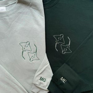 Puede incluir: Dos sudaderas de manga larga, una gris claro y otra verde oscuro. Cada sudadera presenta un dise&ntilde;o bordado de dos rayas manta en hilo verde. La sudadera gris claro tiene "MG" bordado en el pu&ntilde;o, y la verde oscuro "ME".