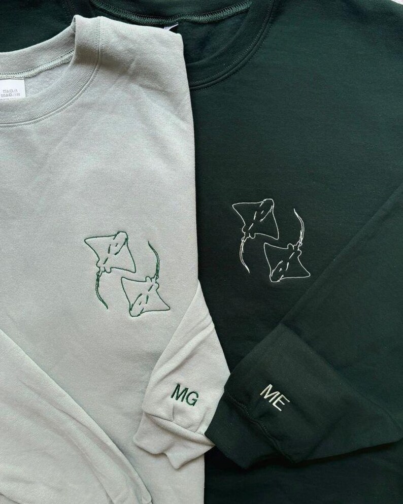 Puede incluir: Dos sudaderas de manga larga, una gris claro y otra verde oscuro. Cada sudadera presenta un dise&ntilde;o bordado de dos rayas manta en hilo verde. La sudadera gris claro tiene "MG" bordado en el pu&ntilde;o, y la verde oscuro "ME".