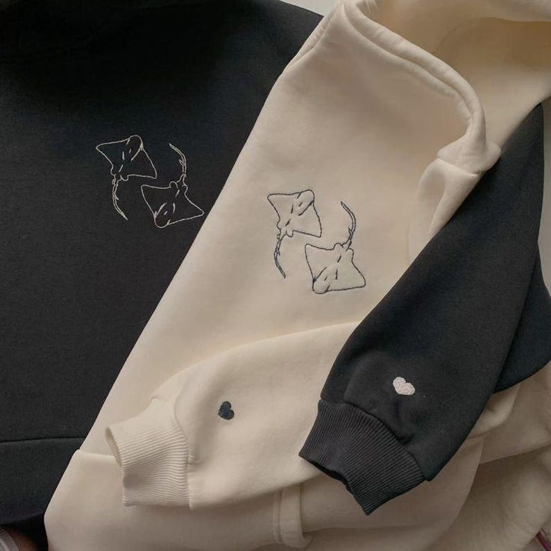 May include: Three sweatshirts in black and cream colors. Each sweatshirt features an embroidered design of two manta rays. The cuffs of the sleeves have a small heart embroidered on them. The sweatshirts are made of a soft material.