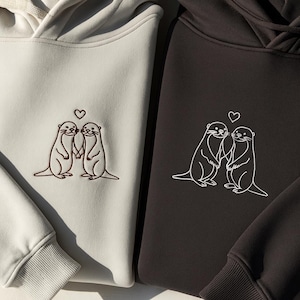 Könnte beinhalten: Zwei Kapuzenpullover, einer hellbeige und einer dunkelgrau, jeweils mit einem gestickten Design von zwei Ottern, die sich unter einem Herzen an den Händen halten. Die Kapuzenpullover haben einen schlichten, minimalistischen Stil.