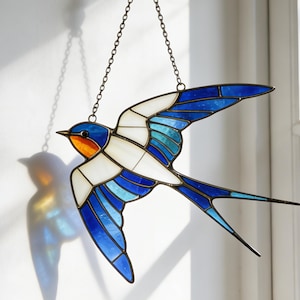 Puede incluir: Atrapasoles de vidriera con forma de golondrina, con detalles azules, blancos y naranjas. El pájaro está suspendido por una cadena y un gancho decorativo. El atrapasoles cuelga frente a una ventana, proyectando una sombra en la pared.