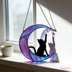 Puede incluir: Una luna creciente de vidriera con una silueta de gato negro que alcanza una estrella. La luna presenta una mezcla de tonos morados y azules y está suspendida por una cadena. Una pequeña planta en maceta se encuentra en el alféizar de la ventana en el fondo.