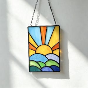 May include: A rectangular stained glass suncatcher featuring a sunrise scene with a yellow sun, orange rays, and blue sky. The lower portion depicts rolling hills in shades of green, blue, and purple. The piece is framed in black metal and hangs from a black chain.