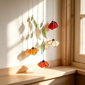 Può includere: Un mobile sospeso in vetro colorato composto da tre fili verticali con foglie in vetro verde e cinque fiori nelle tonalità del rosso, arancione e bianco. Gli elementi sono collegati da sottili fili metallici e una catena centrale delicata.