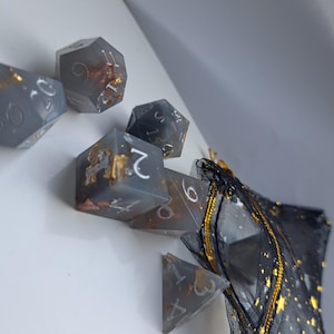 May include: A collection of grey resin dice with gold foil detailing and white numbering. The dice, including a d20, are in various polyhedral shapes. They are presented with a black mesh bag featuring gold star and string accents. Ideal for tabletop games.