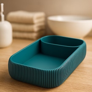 Può includere: Organizzatore da bagno rettangolare color teal con esterno a coste e interno diviso. Un dispenser di sapone bianco, asciugamani, una ciotola e una bottiglia sono sullo sfondo.