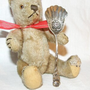 Peut inclure: Un ours en peluche vintage avec un ruban rouge autour du cou tient une cuillère décorative en argent. La cuillère présente un bol orné en forme de coquillage et un motif floral détaillé sur le manche.