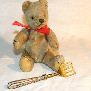 Op de afbeelding: Een vintage teddybeer met een rode strik om zijn nek zit naast een zilveren en goudkleurige serveervork. De beer is lichtbruin met zwarte gestikte details. De vork heeft een decoratief handvat.