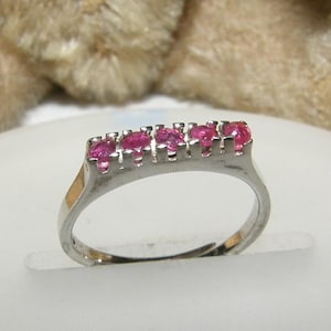 Può includere: Un anello d'argento con cinque vivaci pietre preziose rosa. Le pietre sono disposte in fila sulla parte superiore dell'anello. L'anello è presentato su una superficie bianca, con uno sfondo morbido e sfocato.