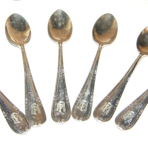 Peut inclure: Six cuillères en argent disposées en éventail. Chaque cuillère a un bol ovale et un long manche avec des gravures décoratives et un monogramme. Les cuillères sont présentées sur fond blanc.