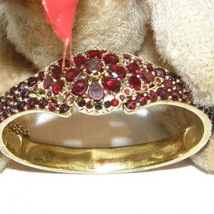 May include: A gold-toned hinged bracelet with a cluster of red gemstones. The bracelet's design features a central section of closely set, oval and round red stones. The bracelet is set against a soft, beige background.